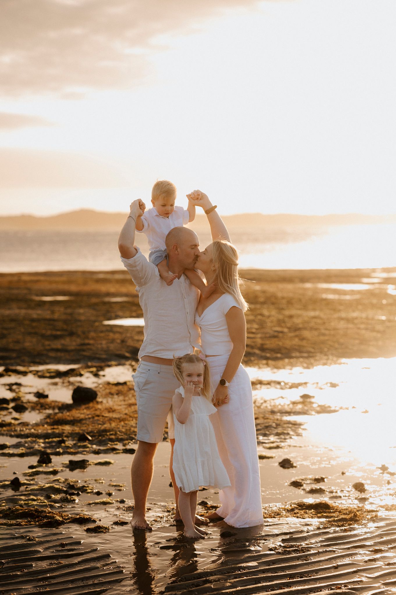 Family of four in white outfits enjoying a golden sunset beach moment, parents kissing while lifting toddler overhead.