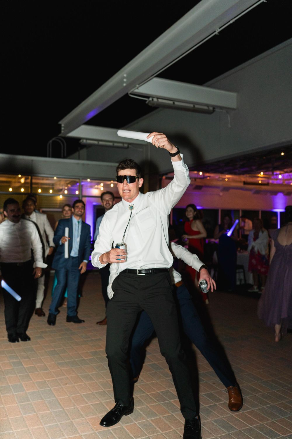 Man in white shirt and sunglasses dancing energetically with a glow stick at an outdoor wedding reception party.