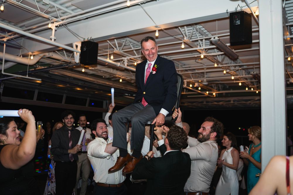 Groom lifted by wedding guests during lively reception celebration on dance floor.