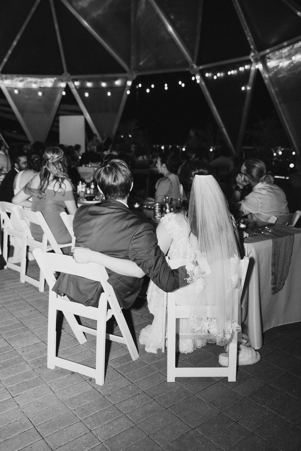 Black and white photo of bride and groom seated at wedding reception table under string lights in elegant dome venue.