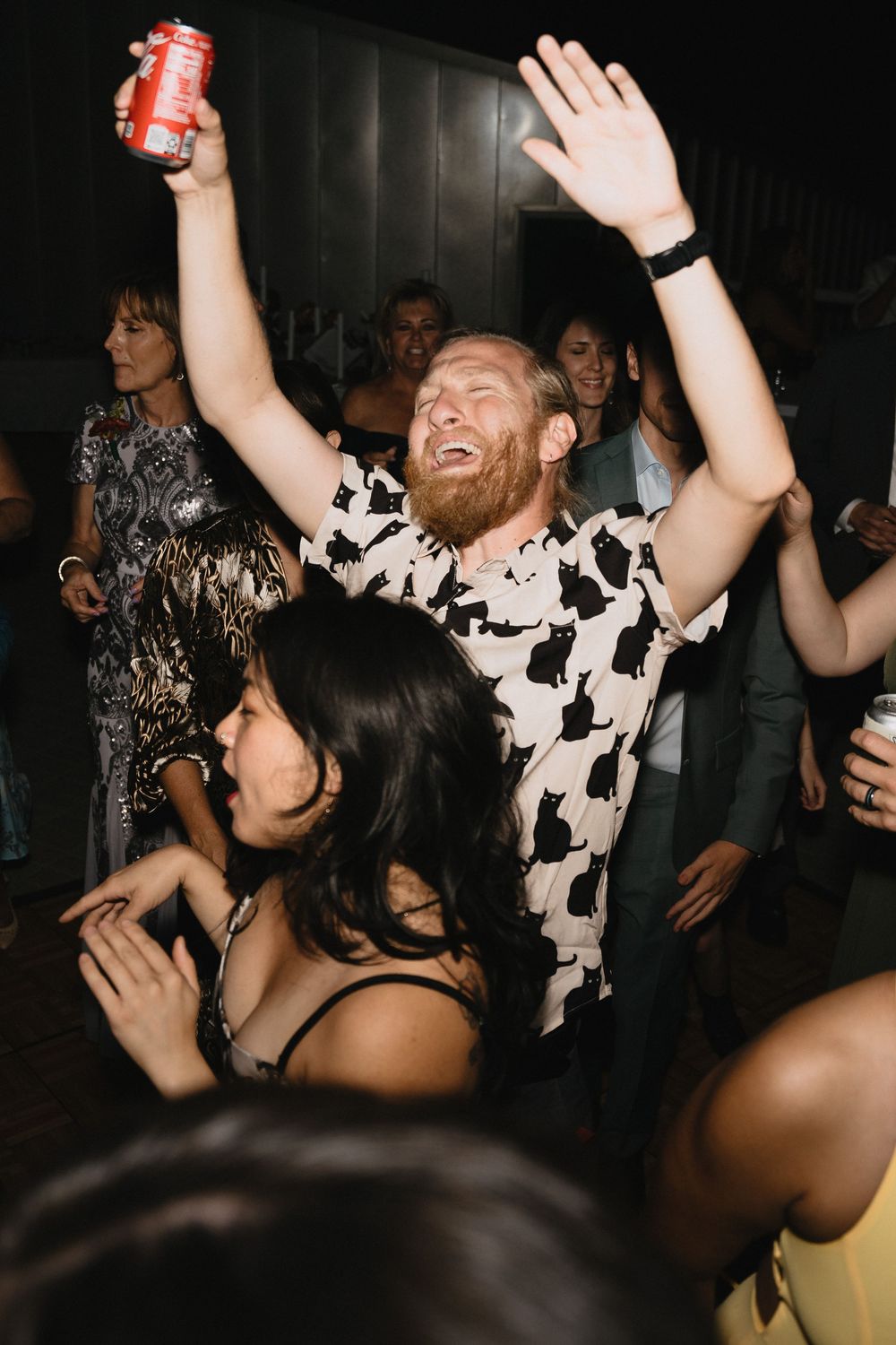 A redheaded man in a cat-print shirt dances joyfully at a party, holding a can aloft with arms raised.