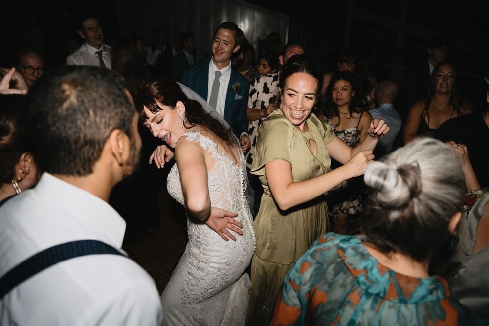 Bride in white lace gown dancing joyfully with guests in elegant attire at a lively wedding reception.