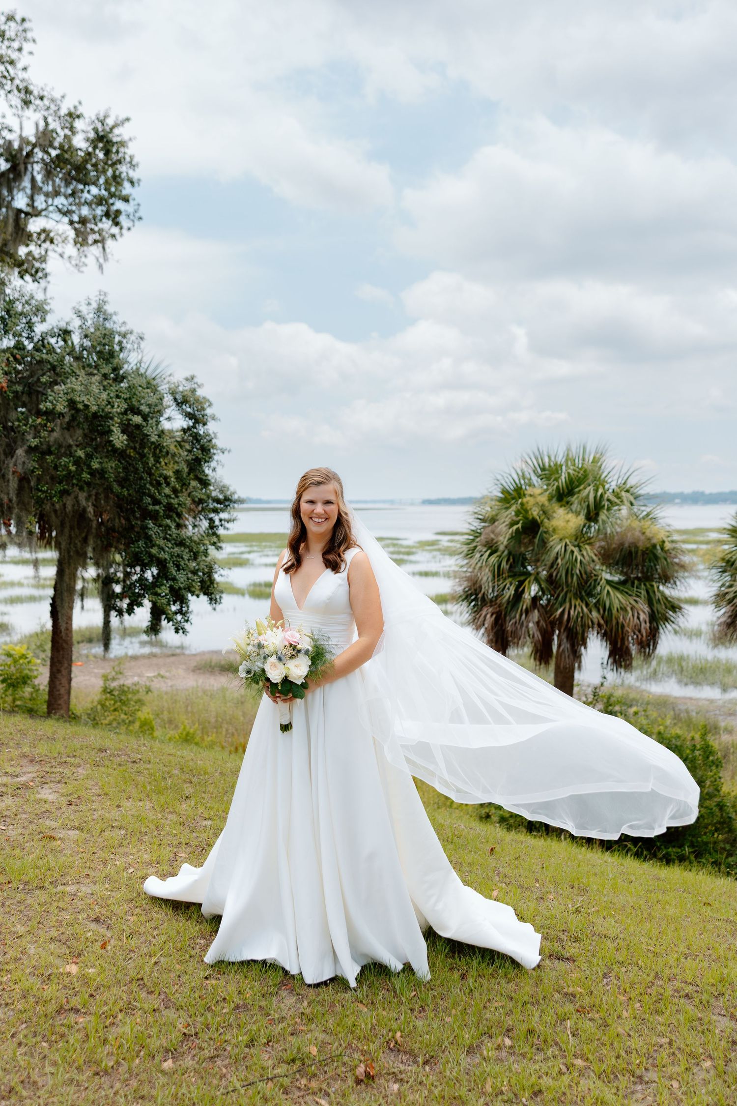 Bride in white ball gown sitting at bay window seat holding white floral bouquet on hardwood floors.