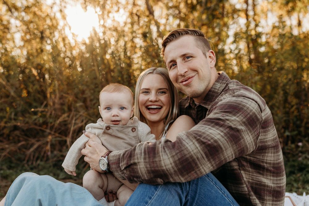 Happy young family with baby boy smiling outdoors in golden autumn light among green trees.