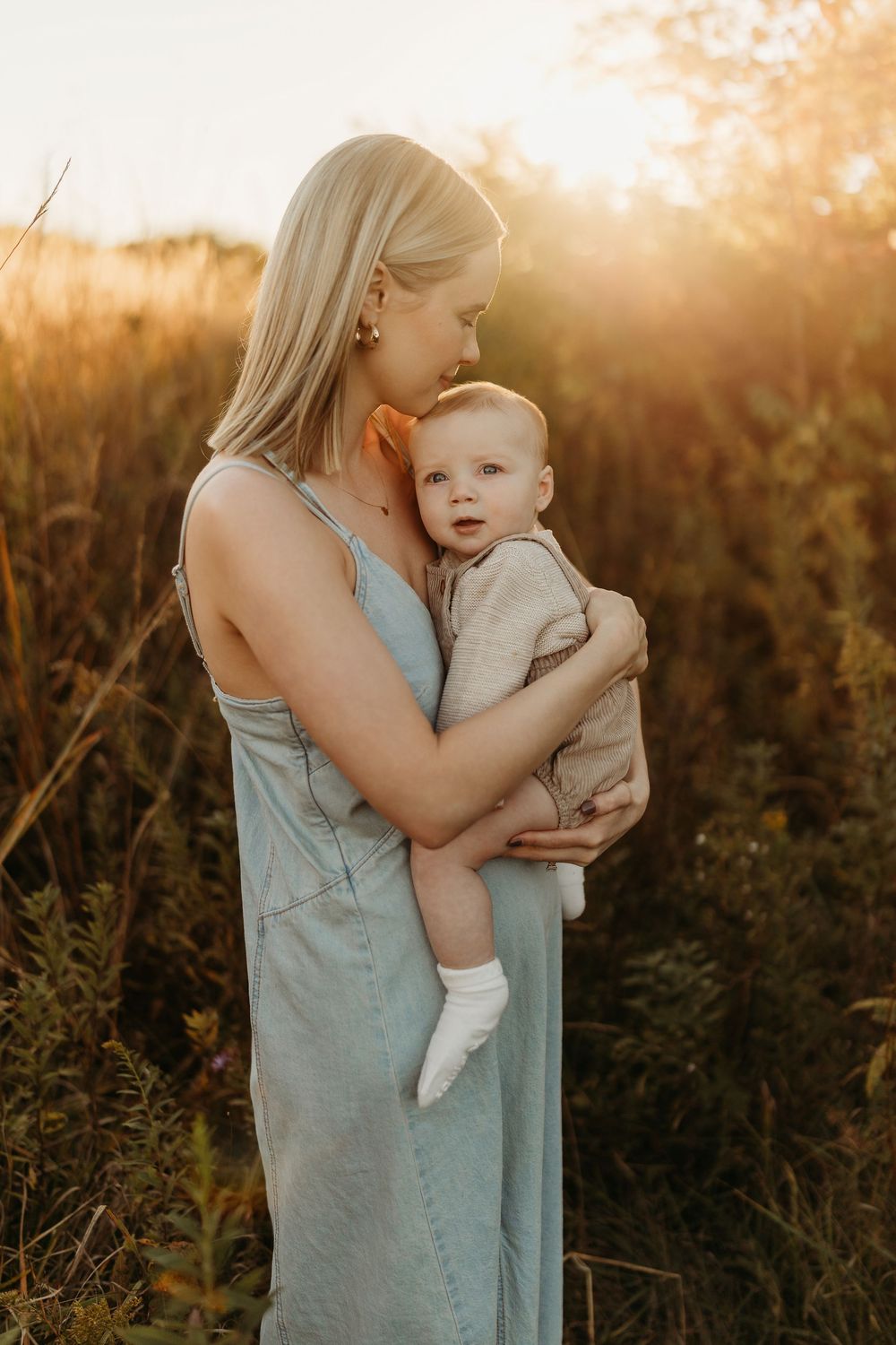 Mother in blue dress holding baby at golden hour sunset in tall grass field.