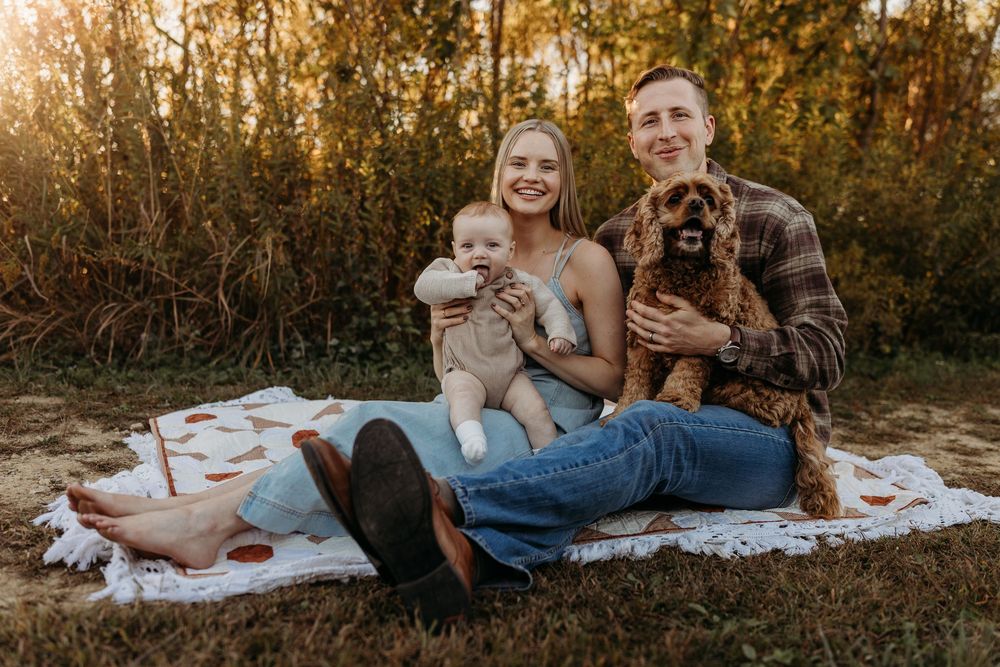 Young family with baby and brown dog sitting on blanket outdoors during golden hour in autumn setting.