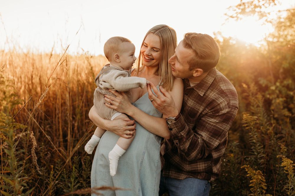 A happy young family with a baby enjoys a golden sunset in a field, the father kissing the mother's cheek.