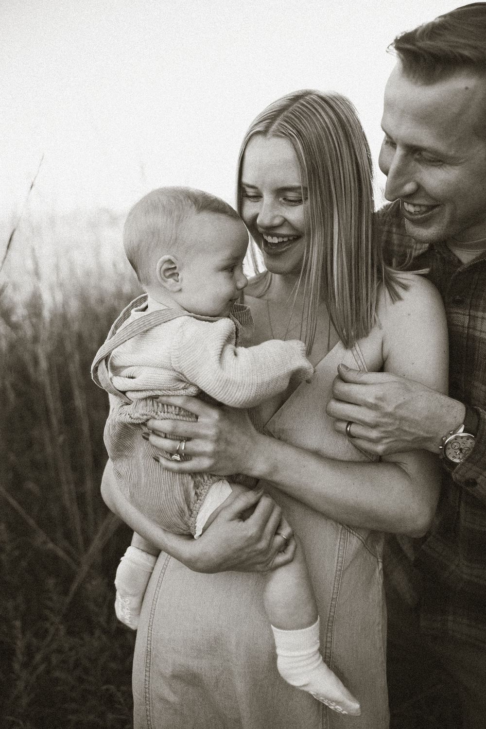 Black and white photo of smiling parents holding their baby outdoors in tall grass.