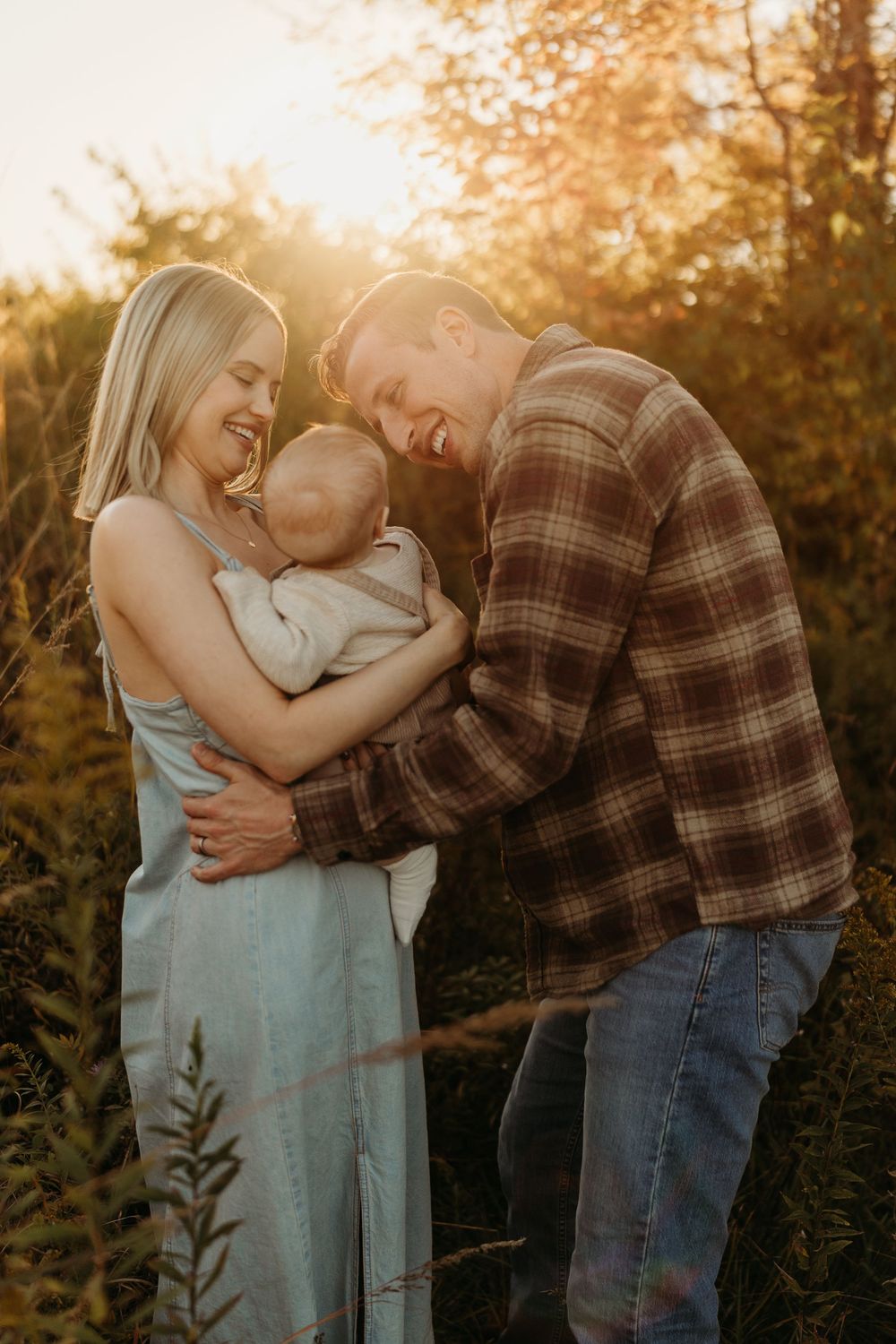 A happy young couple laughs together outdoors at golden hour while cradling their newborn baby in a sunlit forest setting.