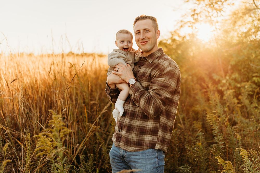A smiling man holds a baby in a sunny golden field, both wearing casual clothes in a warm autumn outdoor setting.