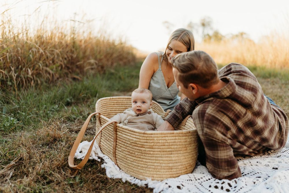 A young family sits on a blanket outdoors, with their baby resting in a large woven basket during a golden hour photo session.
