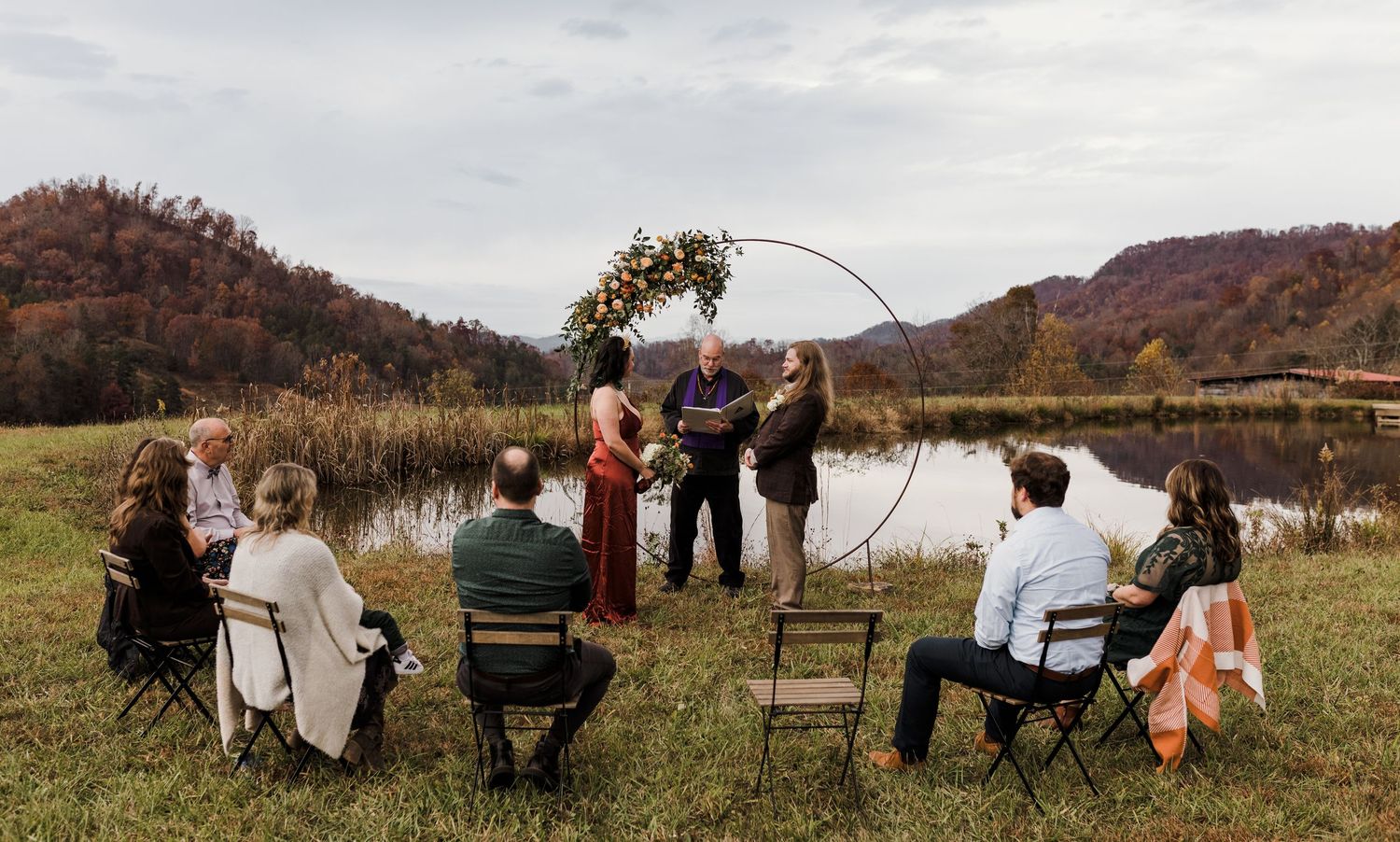 Small intimate outdoor wedding ceremony with circular floral arch by a pond, surrounded by autumn mountain scenery.