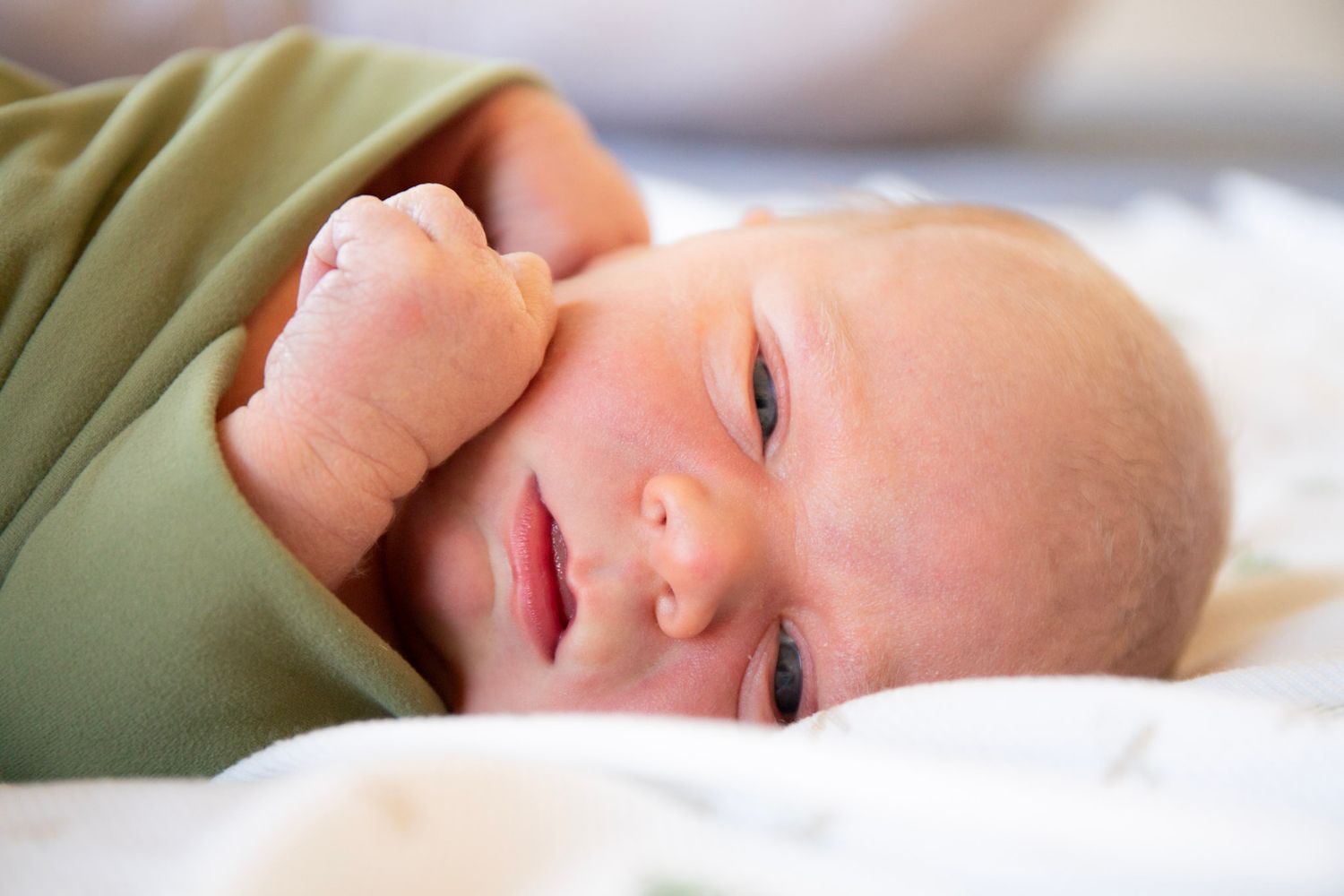 Newborn baby lying on white blanket, wrapped in green swaddle, with tiny fist near cheek.