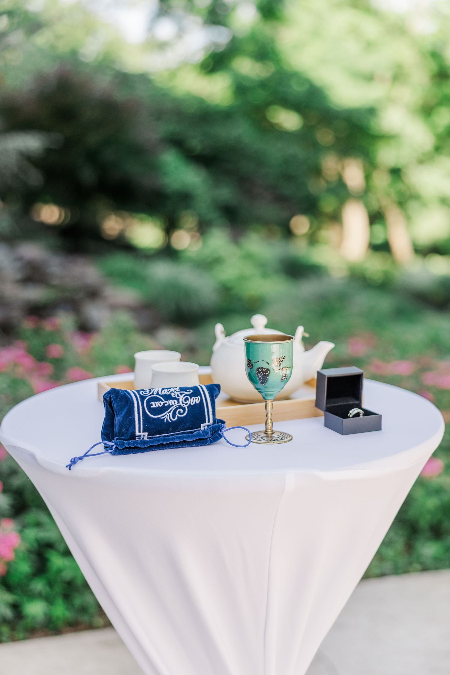 Outdoor wedding ceremony table with kiddush cup, teapot, ring box, and velvet bag on white linen.