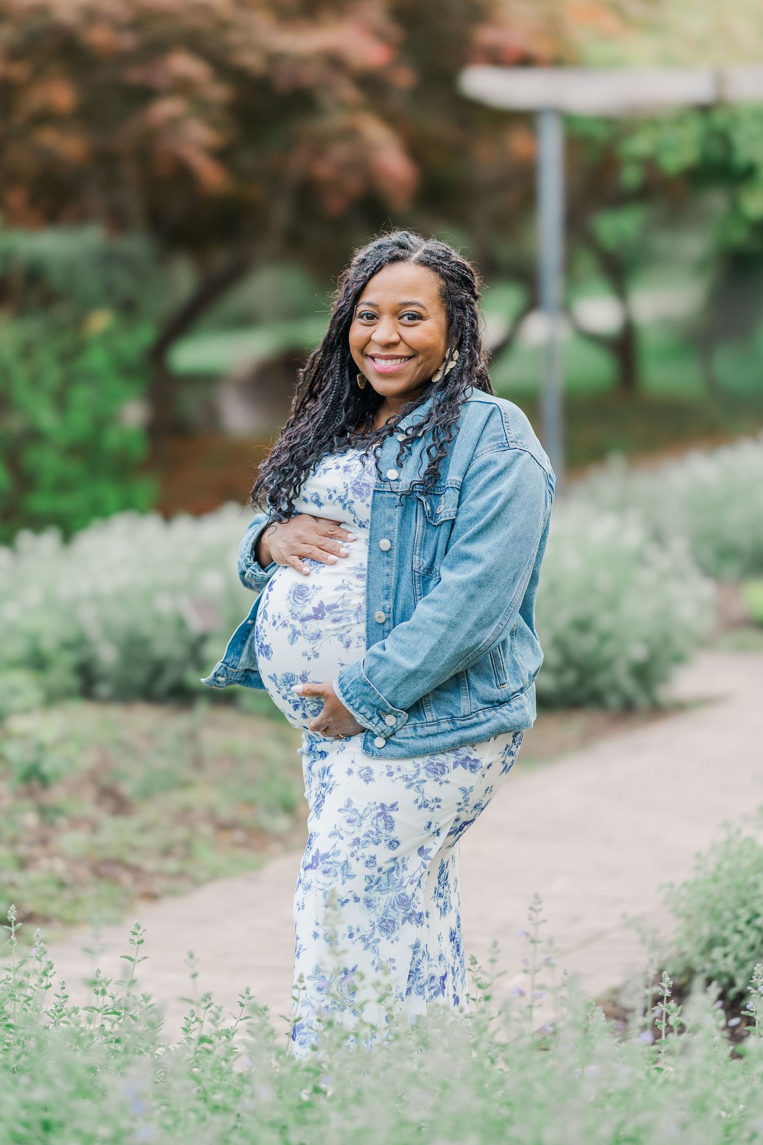 Smiling pregnant woman in floral dress and denim jacket cradling baby bump in a lush garden path.