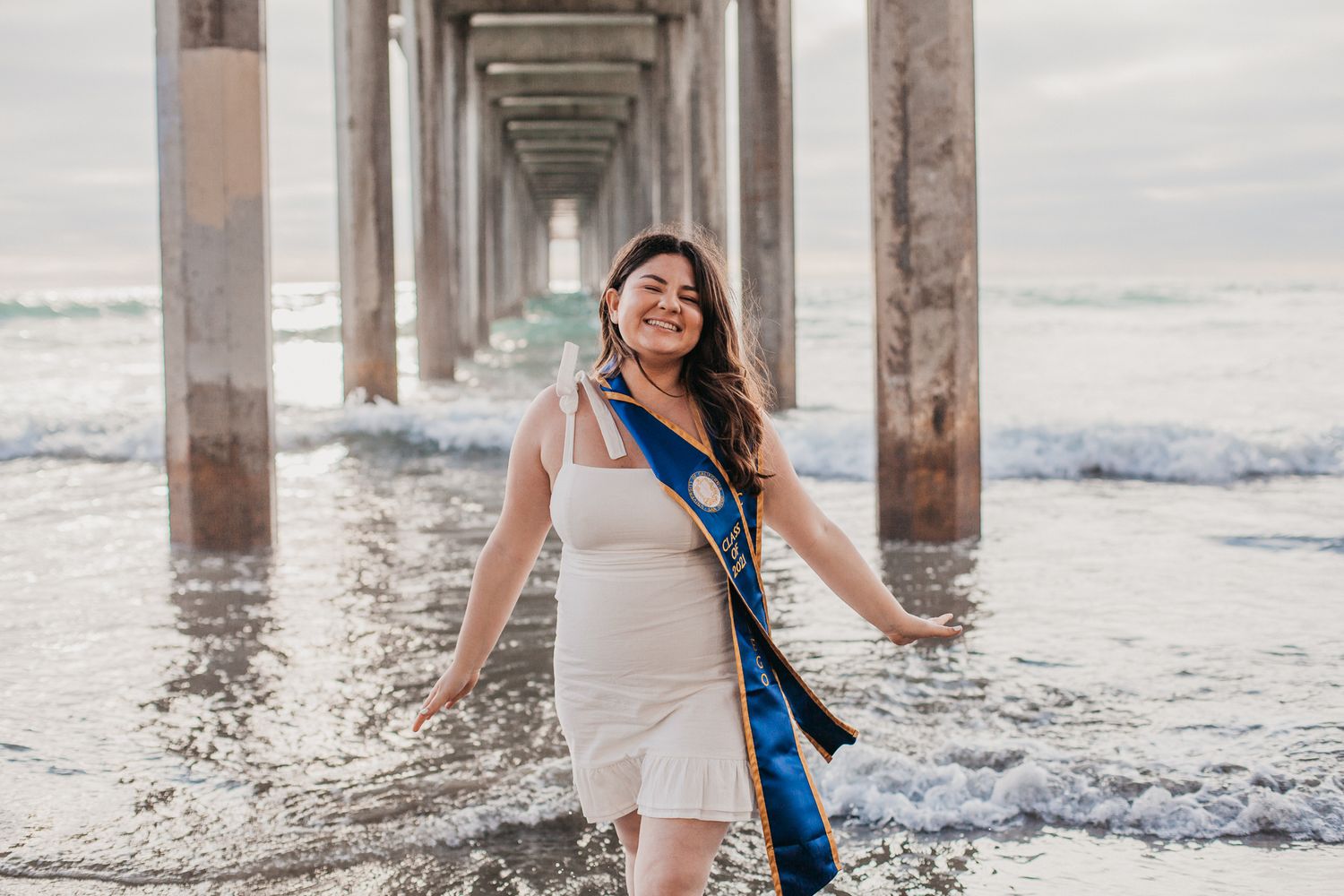 A girl in graduation attire smiles at Scripps Pier with waves in the background.