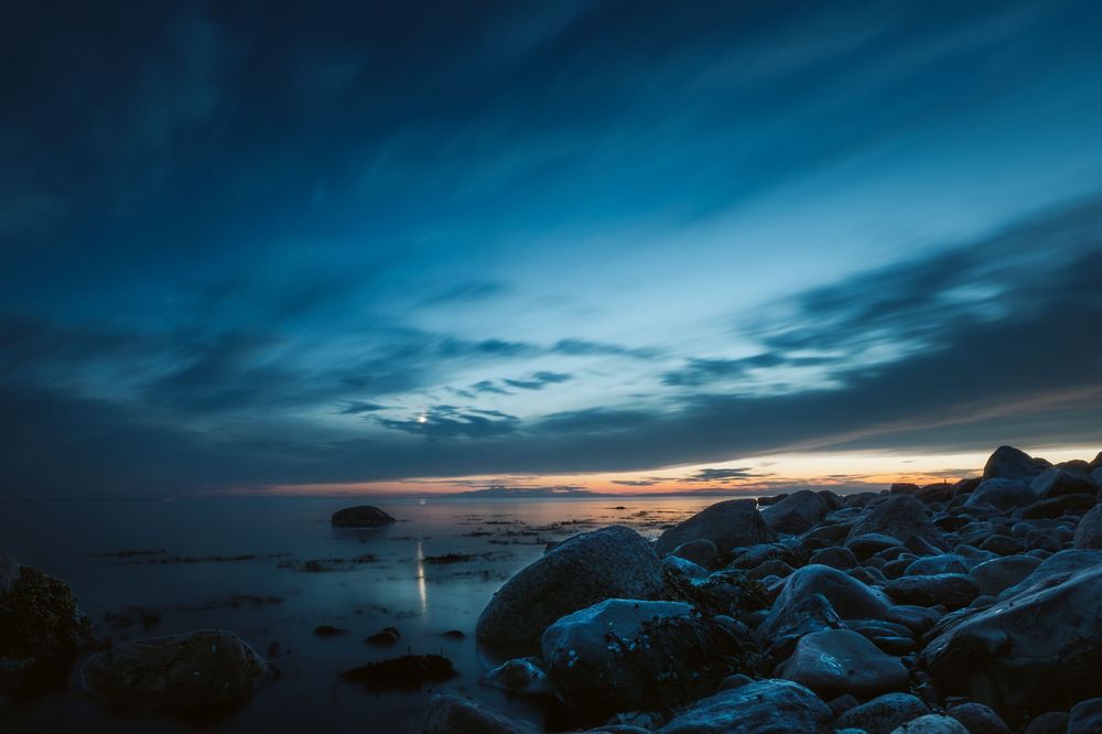A dramatic blue twilight seascape with rocky shoreline and glowing horizon at dusk.