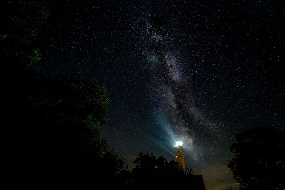 Milky Way galaxy arching over a glowing lighthouse silhouetted by trees under a stunning starry night sky.