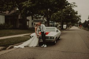 A couple in wedding attire pose next to a vintage white car on a tree-lined residential street.