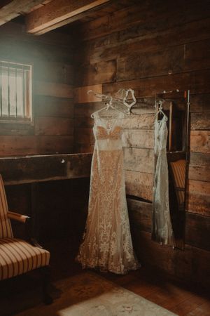 Vintage lace wedding dress hanging in rustic wooden room with natural light.