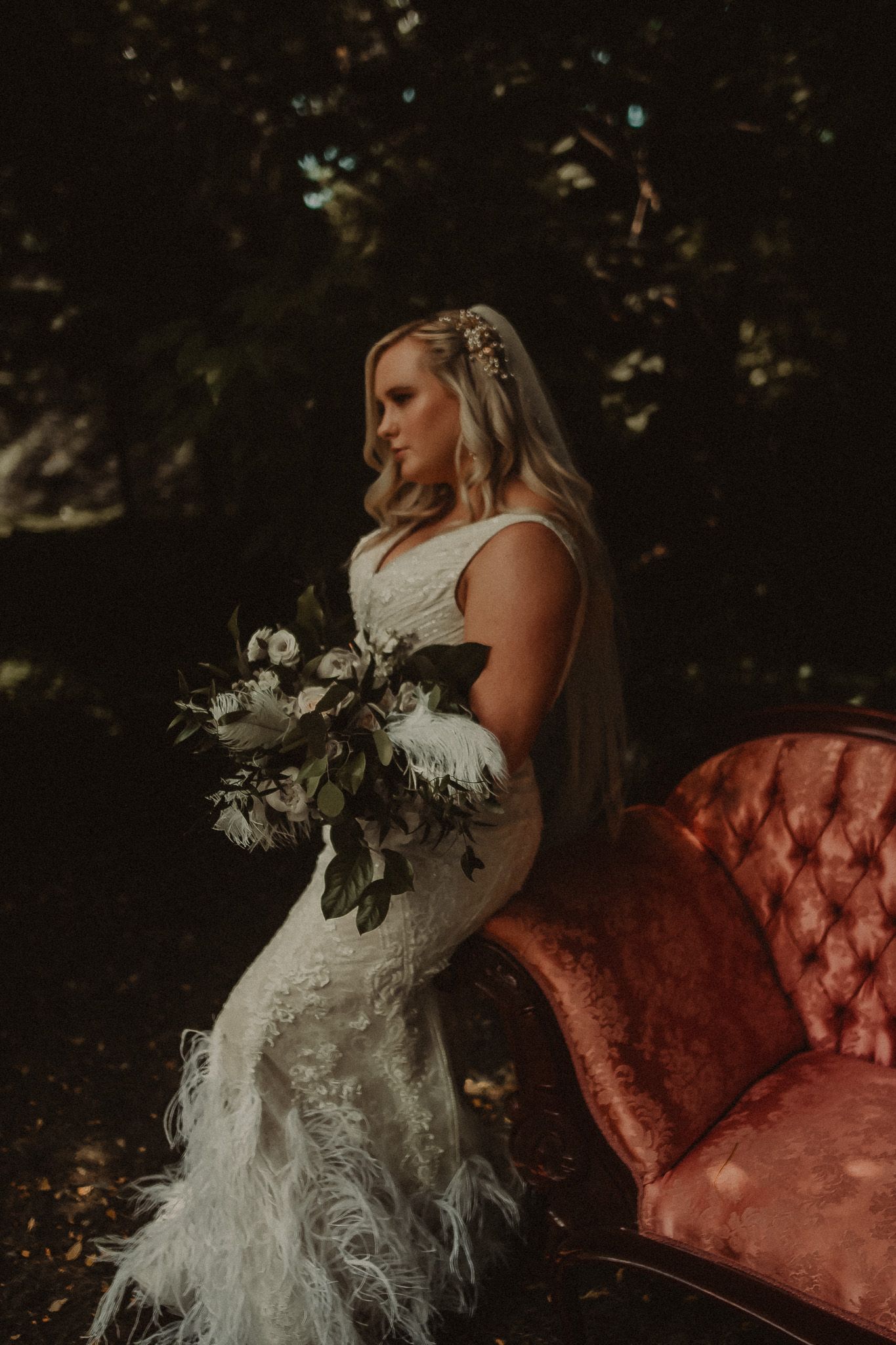 A bride wearing a lace gown holds a white bouquet while posing next to a vintage pink velvet couch in a garden setting.