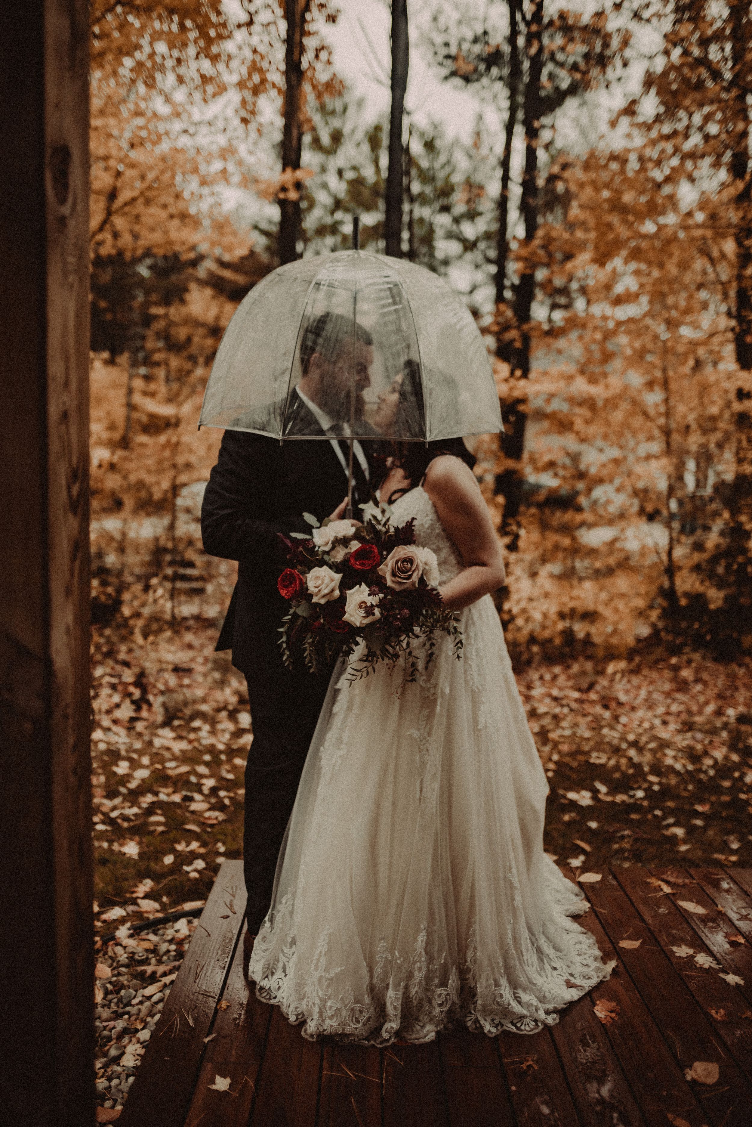 Newlyweds share an intimate moment under a clear umbrella surrounded by autumn foliage on a wooden deck.