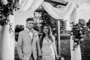 A wedding couple stands under a decorated arch with white draping and floral arrangements.