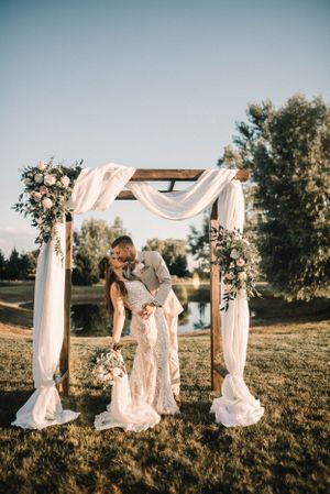 A couple embraces under a wooden wedding arch decorated with white fabric and flowers at sunset.
