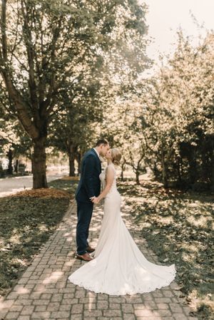 A wedding couple shares an intimate moment on a brick pathway surrounded by sunlit trees.