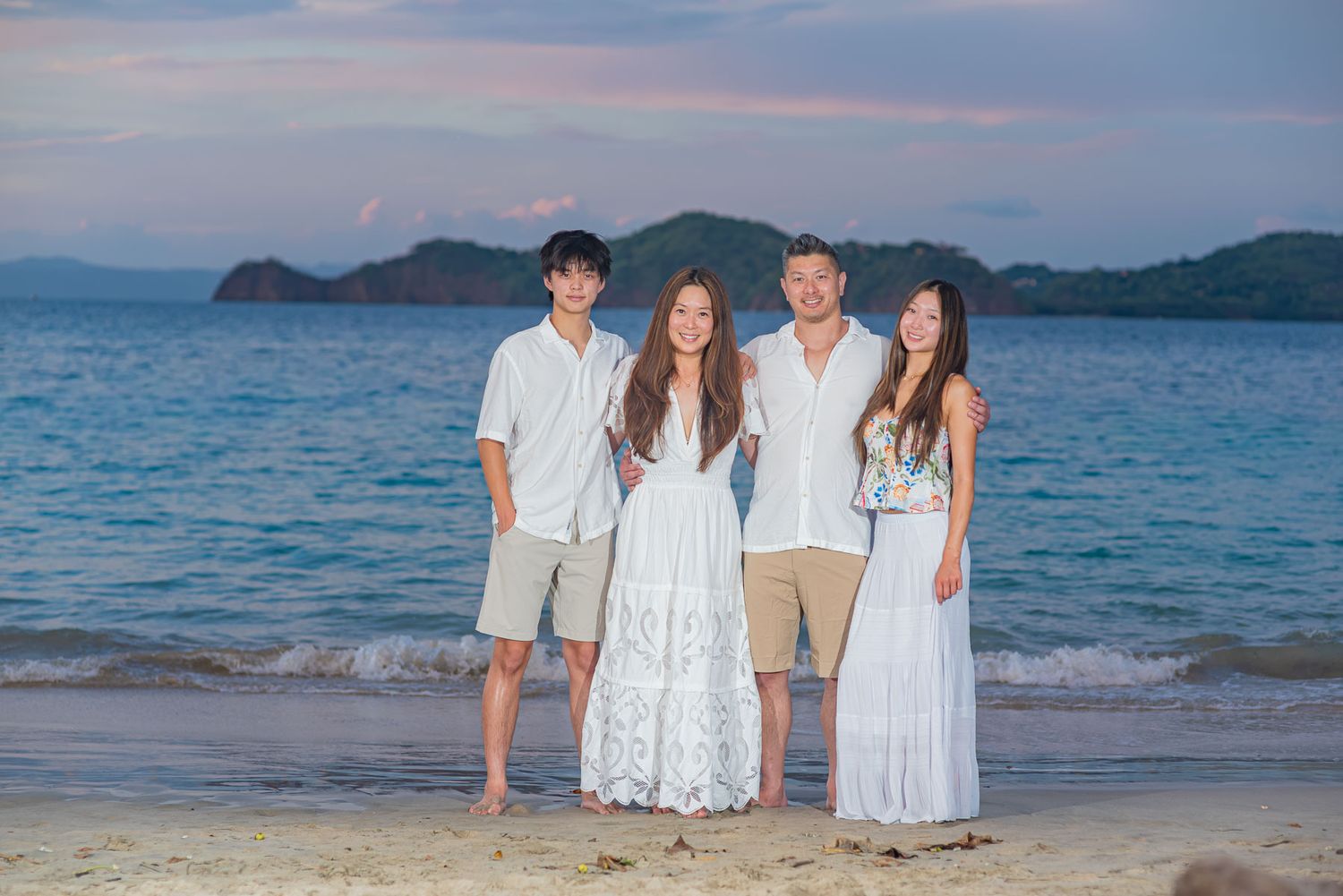 Family of four posing on a sandy beach at sunset with turquoise ocean and tropical islands in the background.