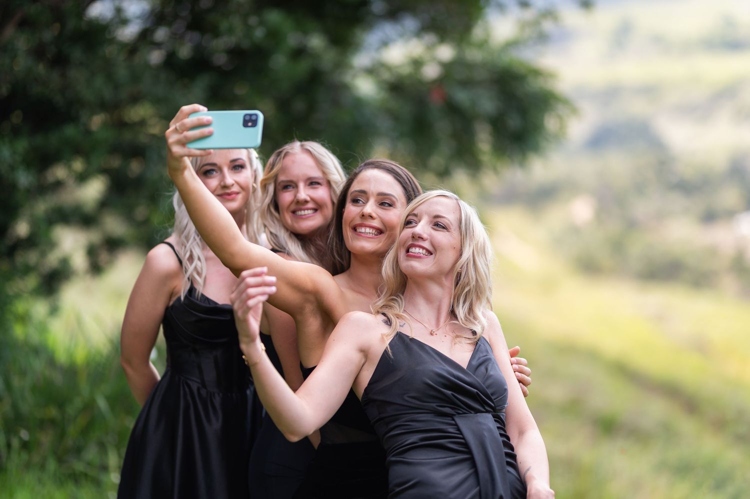 Four women in black dresses smiling and taking a selfie outdoors with green hills in the background.