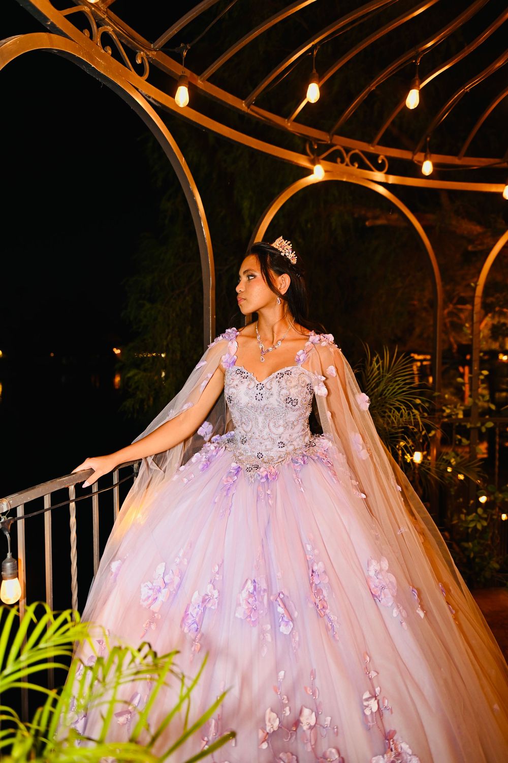 Young woman in a pink floral ball gown and tiara poses gracefully under a lit gazebo at night.