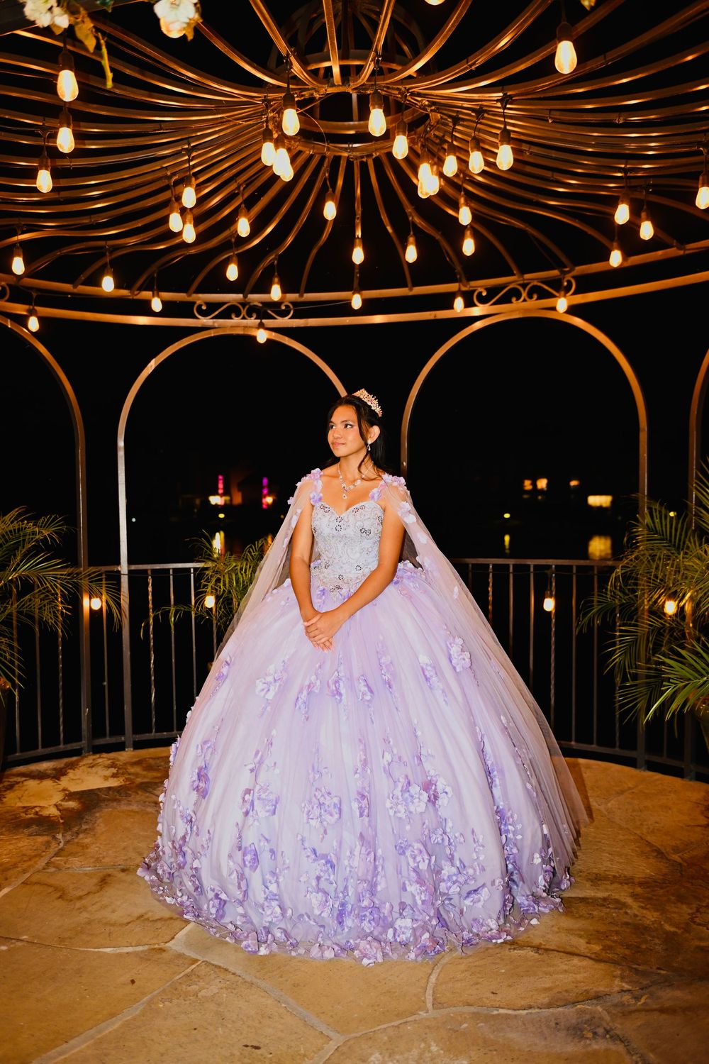 Young woman in lavender quinceañera ball gown stands under ornate gazebo with warm string lights at night.