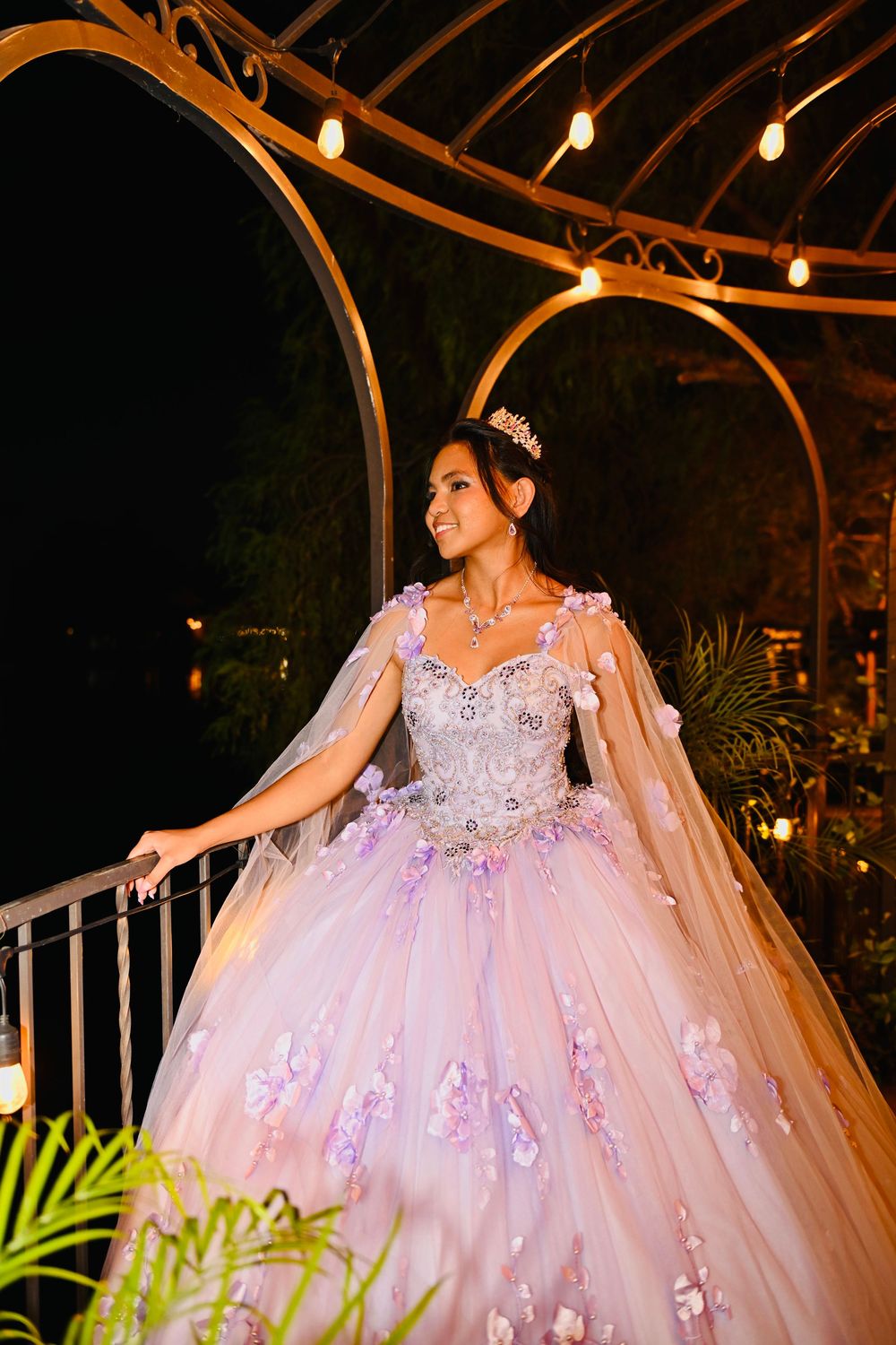 Young woman in a pink floral ball gown and tiara poses under a gazebo with string lights at night.