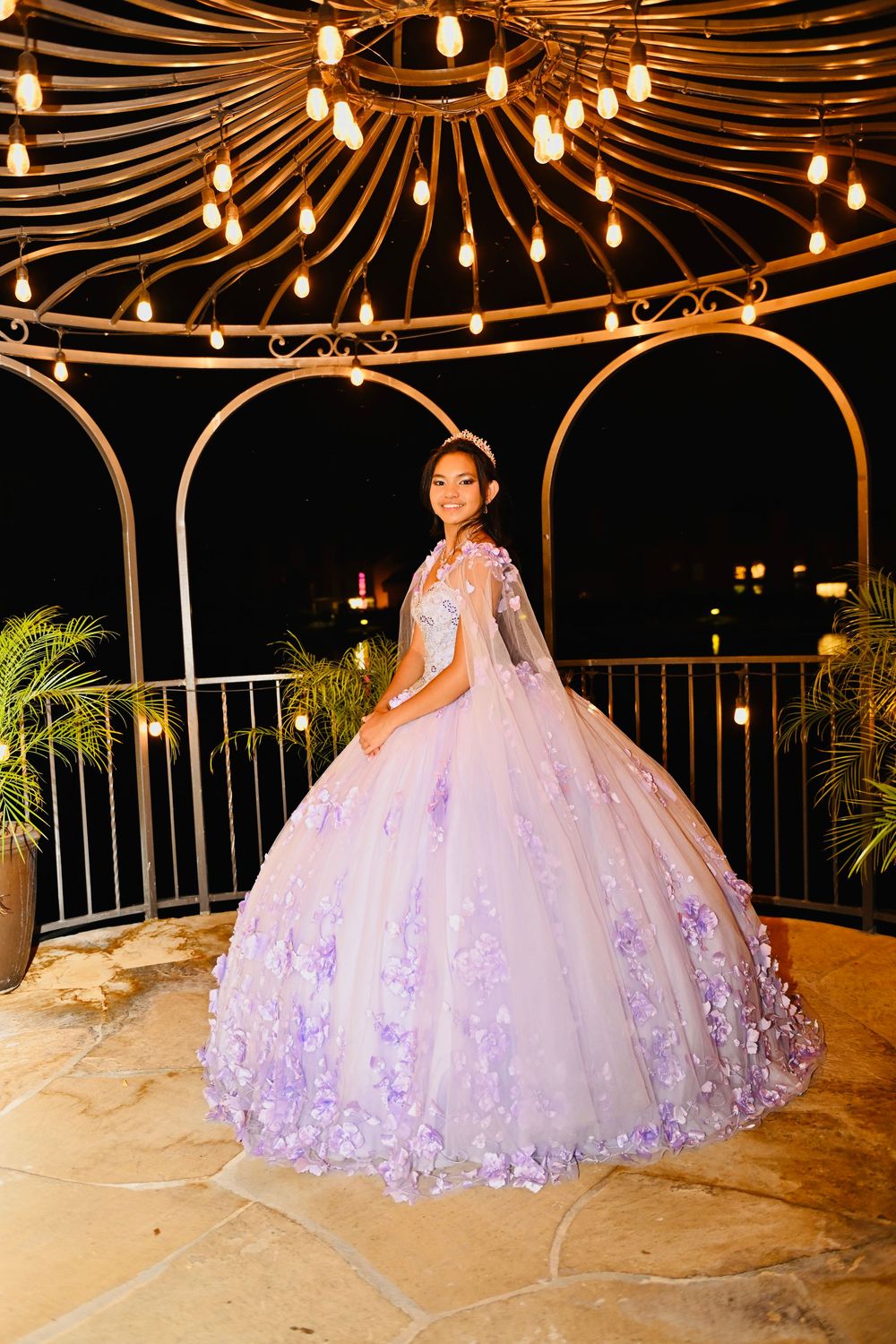 Young woman in lavender quinceañera ball gown with floral appliqués poses under ornate gazebo with warm string lights at night.