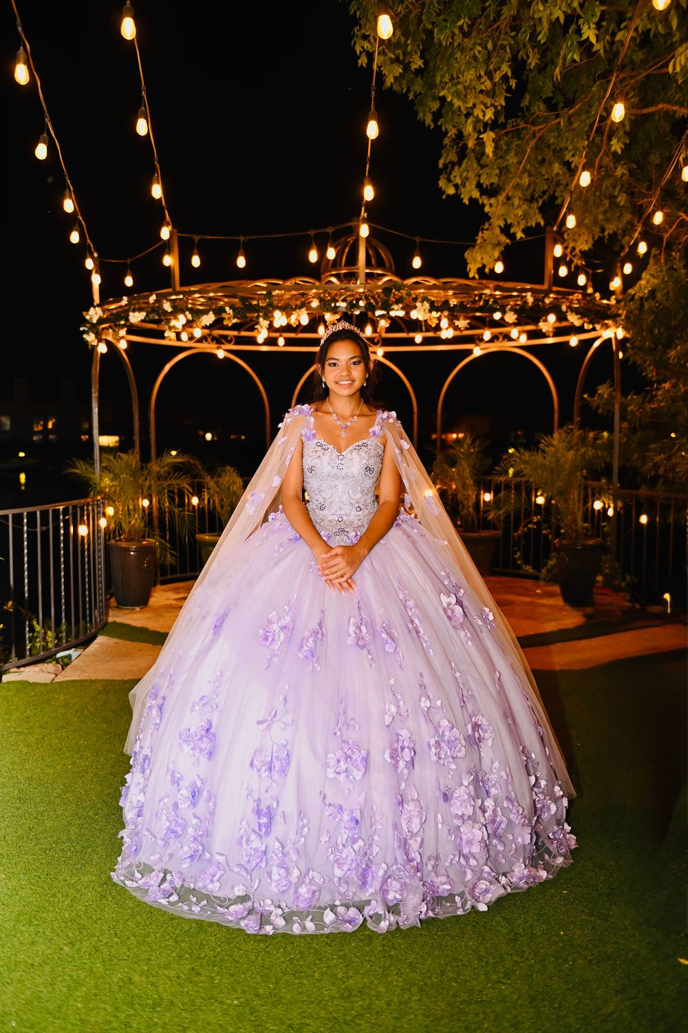 Young woman in lavender ball gown with floral details posing under string lights at quinceañera.