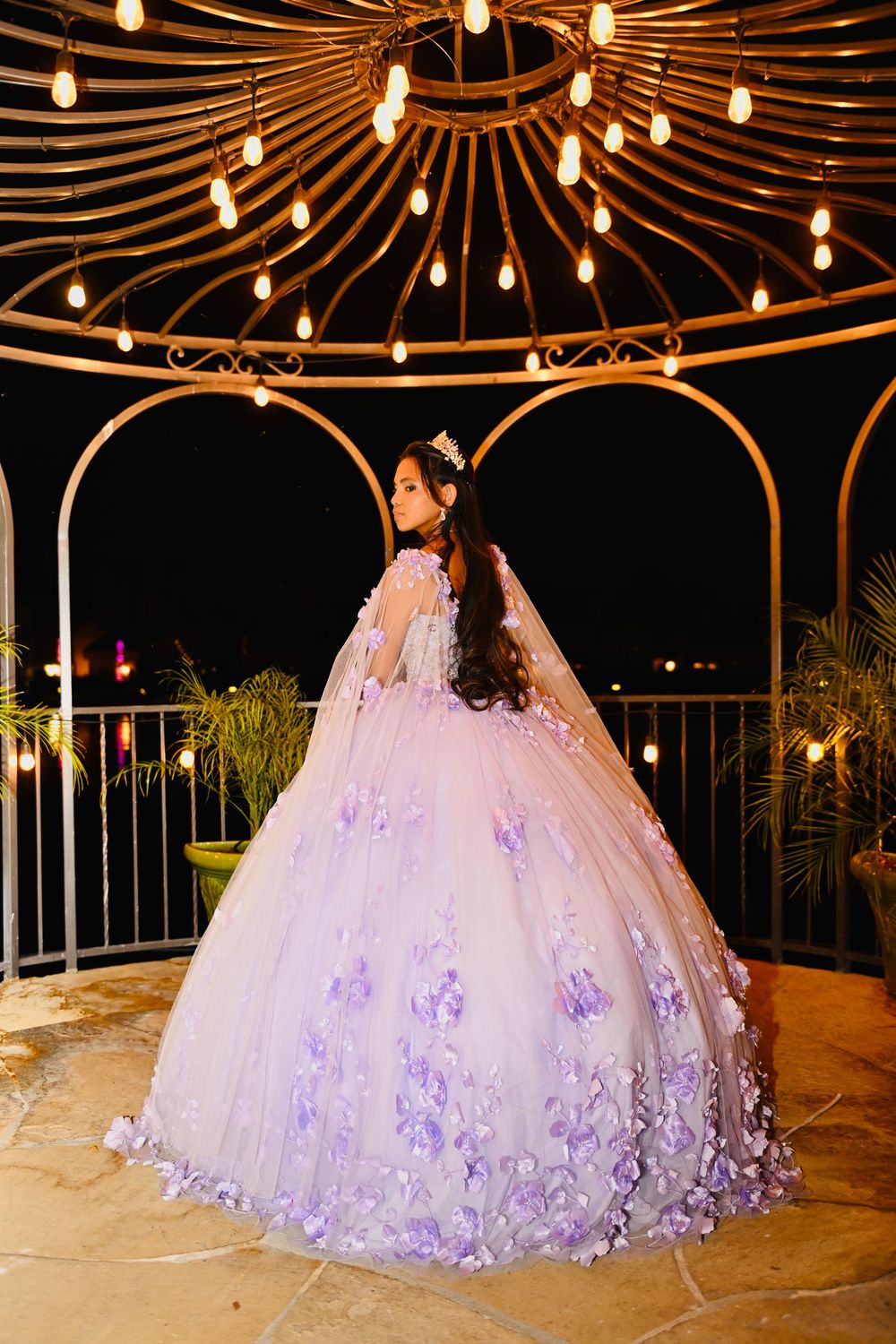 Young woman in lavender quinceañera ball gown with floral details and cape, posing in lit gazebo at night.