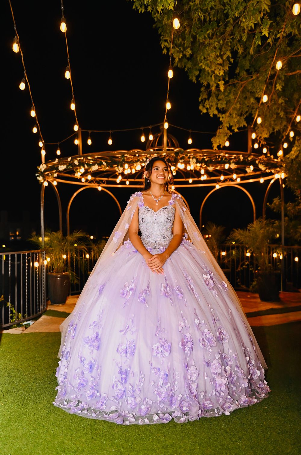 Young woman in lavender ball gown with floral appliques poses under string lights at elegant outdoor quinceañera.