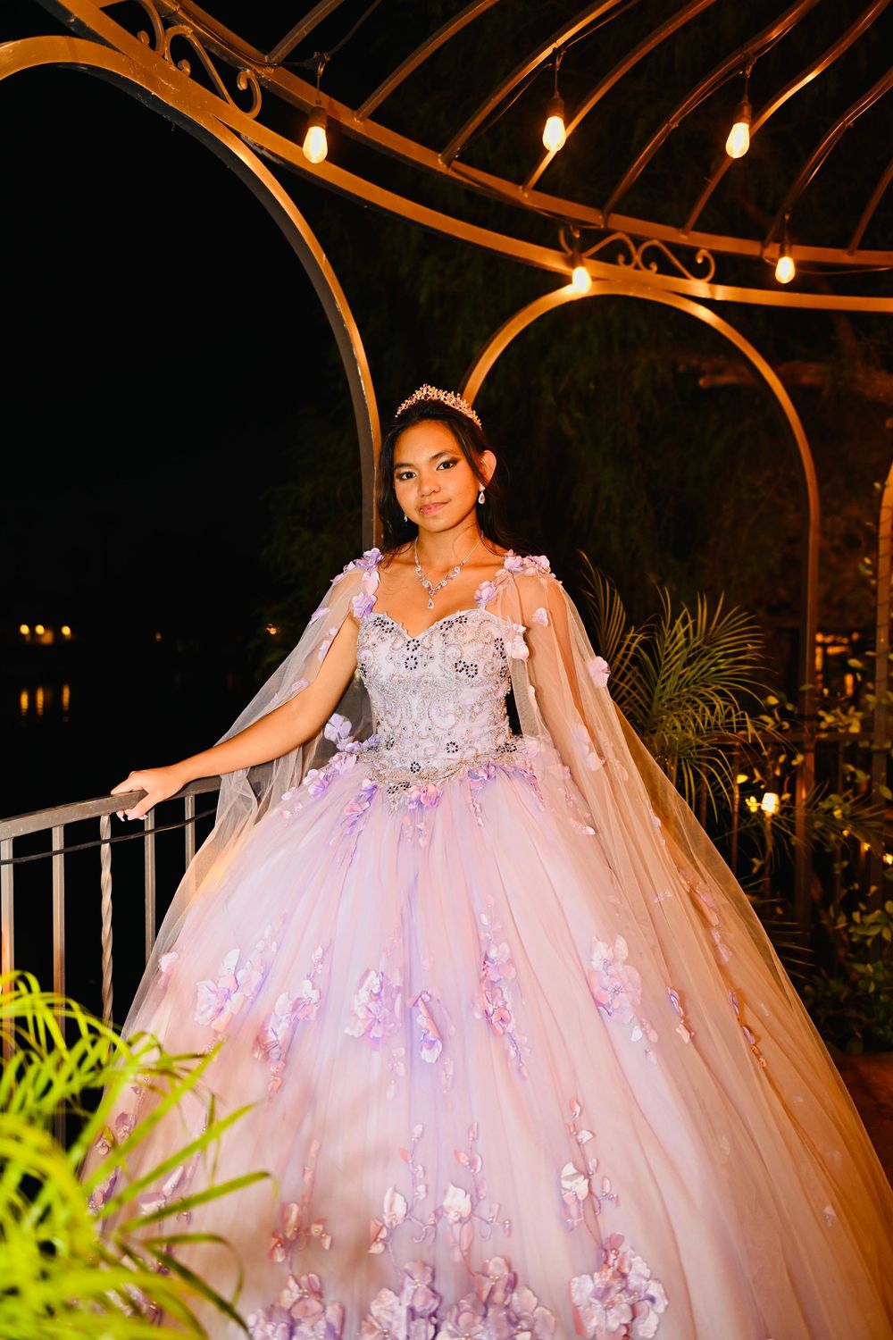 Teen in pink-lavender quinceañera gown with cape sleeves and tiara posing in lit gazebo at night.