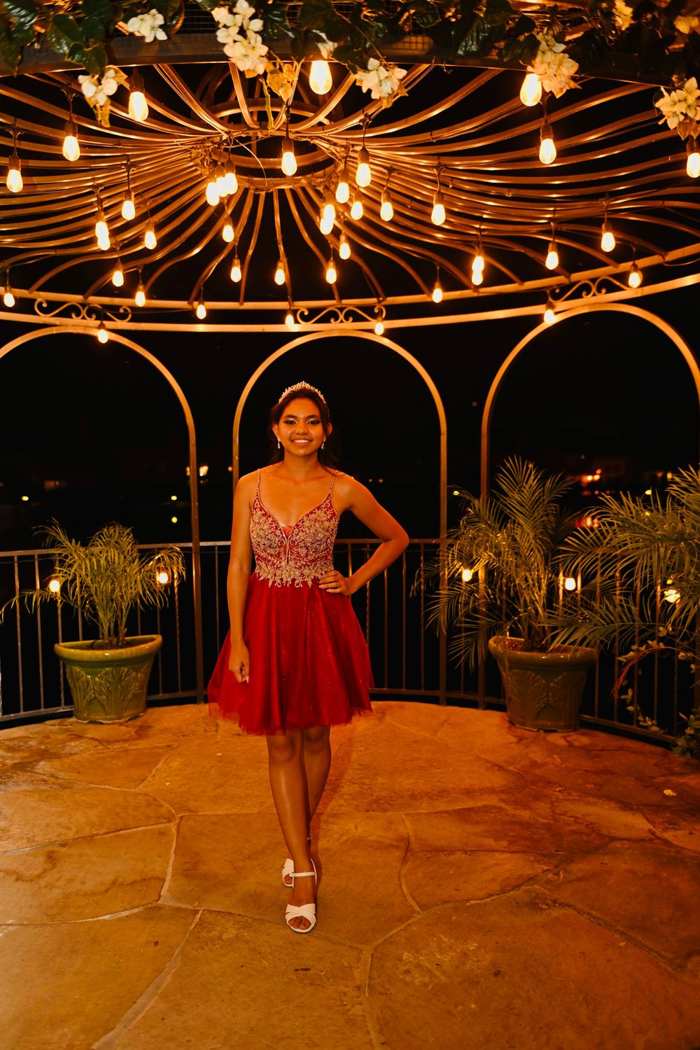 Young woman in red dress poses under ornate gazebo with warm string lights at night.