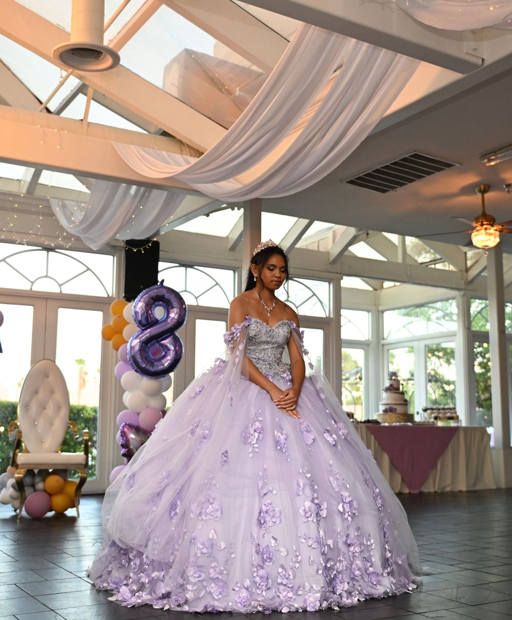 Young woman in lavender ball gown with floral appliques poses at quinceañera celebration in elegant venue.
