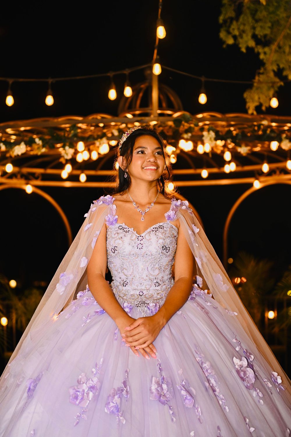 Smiling young woman in lavender quinceañera gown with cape and floral details stands under glowing gazebo string lights at night.