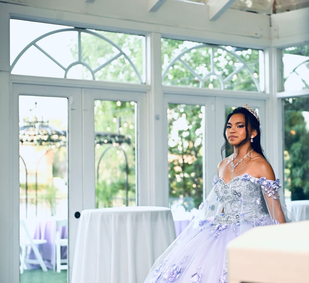 Young woman in lavender off-shoulder ball gown with floral appliques poses in elegant white conservatory.