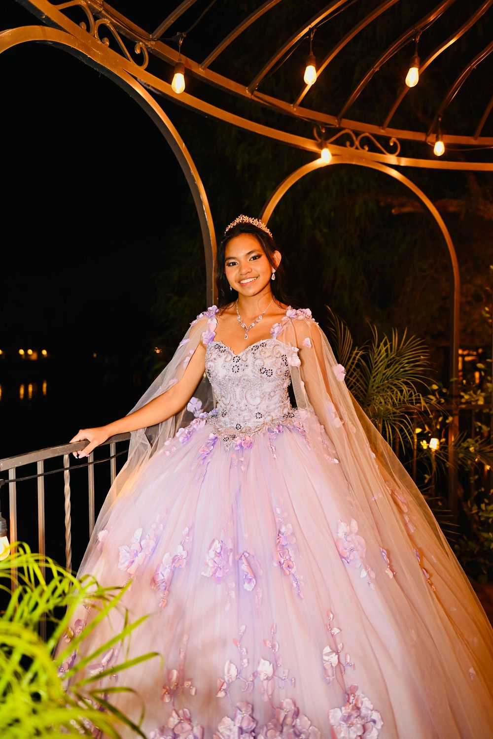 Joyful young woman in lavender quinceañera ball gown with 3D floral appliqués poses gracefully under lit gazebo at night.