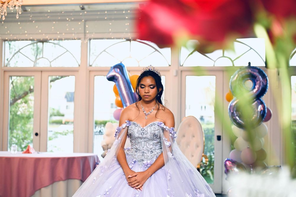 Young woman in lavish silver quinceañera ball gown poses elegantly at decorated venue with number balloons.