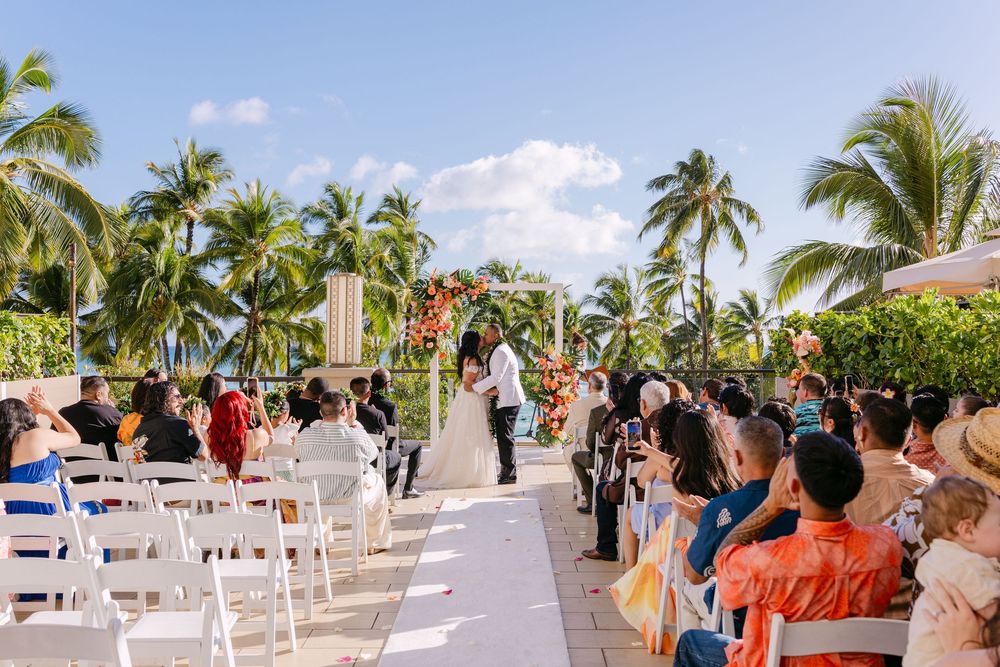 Outdoor tropical wedding ceremony with guests seated in white chairs, palm trees, and couple at the altar under blue skies.