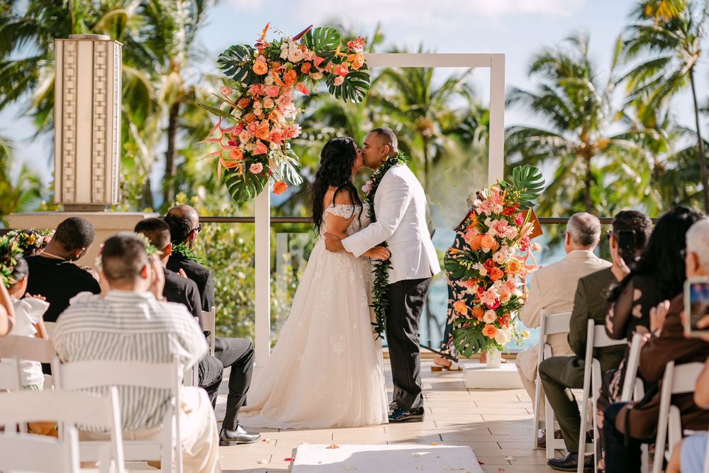 Couple exchanges vows at tropical outdoor wedding ceremony with lush coral floral arch and palm trees in background.