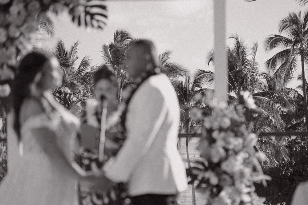 Black and white wedding ceremony photo of couple exchanging vows outdoors with tropical palm trees and floral arrangements.