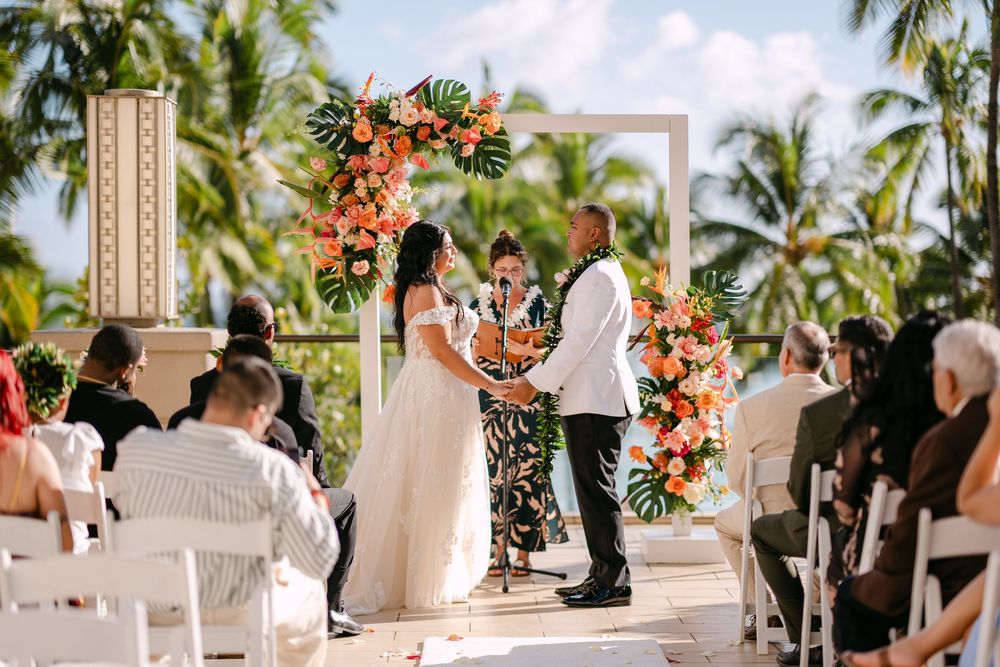 Couple exchanges vows at tropical outdoor wedding ceremony with lush floral arch and palm trees in background.