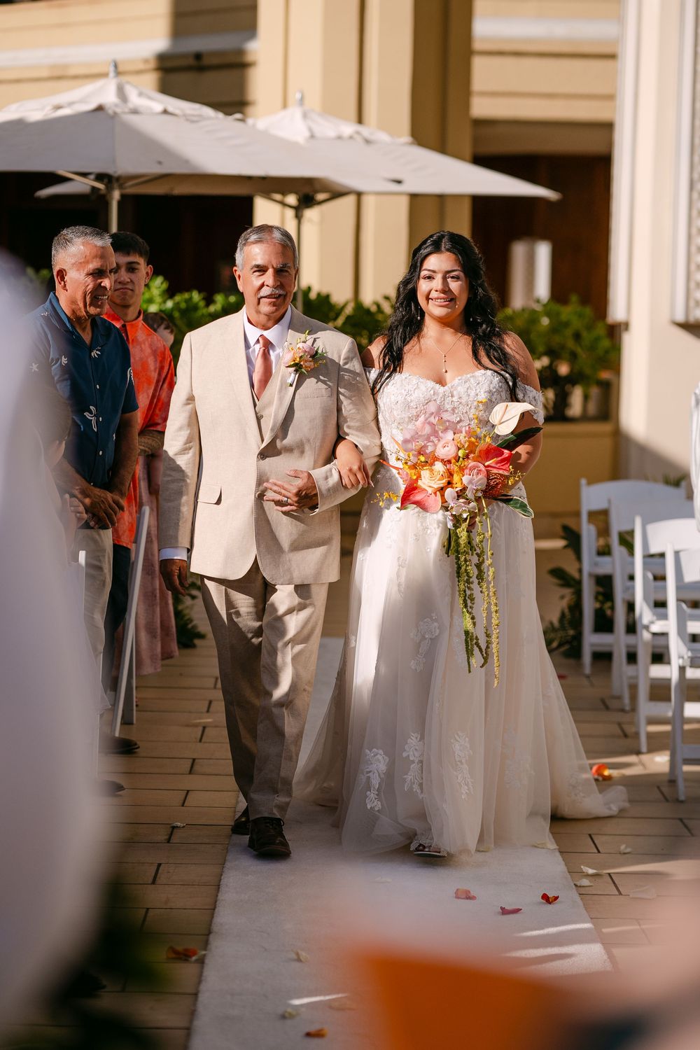Bride walks down outdoor aisle with escort, holding colorful bouquet, in elegant white gown at sunny wedding ceremony.